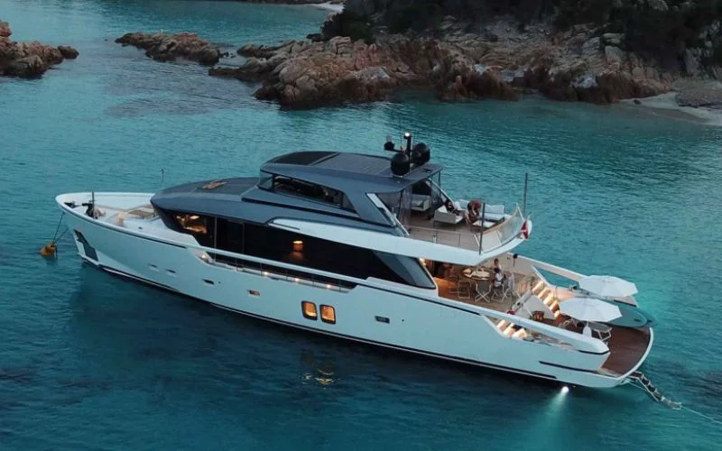 Beautiful shot of the yacht Tiberias by Malayachts.ae, sparkling under lights in a tranquil, rocky bay at dusk.