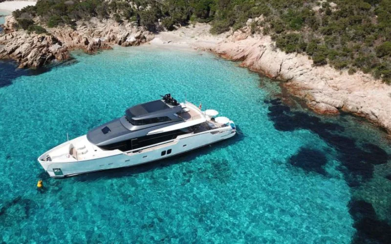 Aerial view of the yacht Tiberias by Malayachts.ae, floating in incredibly clear, vibrant blue-green water surrounded by a lush, rocky coastline.