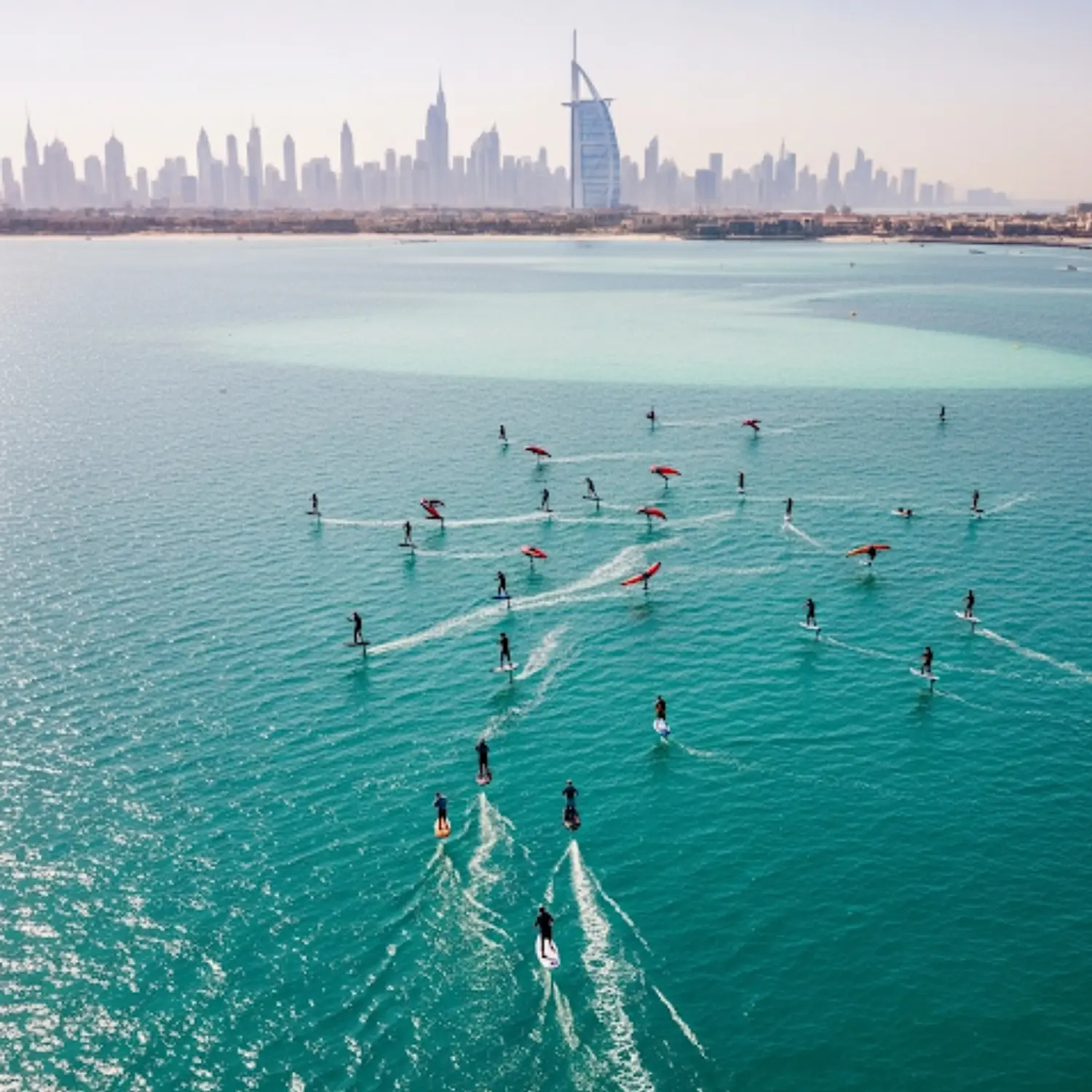 An aerial view shows a large group of people riding electric hydrofoil surfboards on the turquoise ocean. The sail-shaped Burj Al Arab hotel and the Dubai city skyline are visible in the distance on the coast
