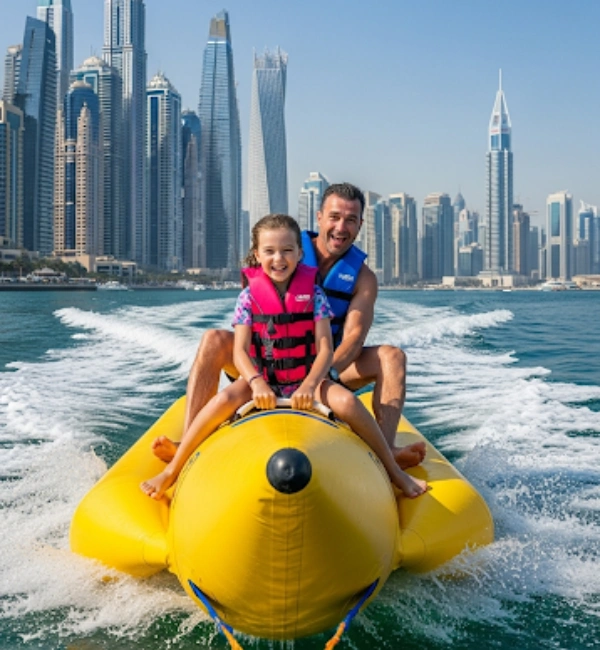 A man and a young girl are sitting on a yellow banana boat, both smiling and wearing life vests. The girl is in front wearing a pink vest, and the man is behind her wearing a blue vest. They are being towed on the water, creating a white wake, with the dense, modern skyline of Dubai in the background.