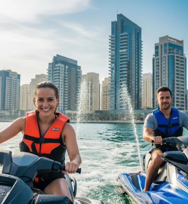A man and a woman are riding separate jet skis on the ocean, both laughing excitedly. The man is in the foreground, wearing sunglasses and a blue life vest. The woman is behind him, wearing an orange life vest and a sun visor. A jet of water shoots into the air between them. In the background, the sail-shaped Burj Al Arab hotel and the Dubai city skyline are visible under a clear, bright sky.